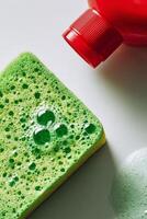 Bright Green Sponge and Red Soap Bottle With Bubbles on a White Surface During a Cleaning Session. photo