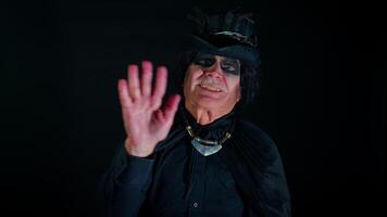 Frightening senior man with Halloween witcher makeup waves hand palm in hello gesture welcomes someone with hospitable expression expresses positive emotions. Sinister guy on studio black background video
