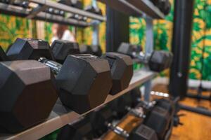 A rack of hexagonal dumbbells in a gym with tropical themed wallpaper, wooden flooring, and a blurred figure in white in the background. photo