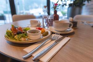 A dining table set with salad, smoked salmon, olives, coffee, and water. Adorned with fine cutlery and a red flower, with serene outdoor views. photo
