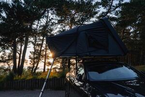 A modern SUV with a dark rooftop tent and ladder is parked in a forest. Tall trees surround the vehicle, with sunlight glowing through the canopy. A body of water is visible in the distance. photo