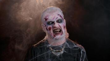 Zombie man with makeup with fake wounds scars and white contact lenses looking at camera and clicks his teeth, trying to scare in dark room. Sinister dead guy. Halloween, filming, staging concept video