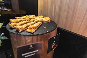 Display of sliced pastries with crumbly topping on a black slate platter atop a wooden stand, set against a wooden panel and black countertop. photo