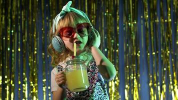 Futuristic hipster teen girl listening to music in headphones, dancing in slow motion at disco party cyberpunk club. Child kid in neon lights with pineapple fruit juice standing by illuminated wall video