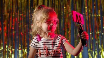 Trendy stylish child kid blogger smiling cheerfully, recording blog vlog on smartphone with tripod. Fashion girl in futuristic night club with red neon lights making selfie call on mobile phone video