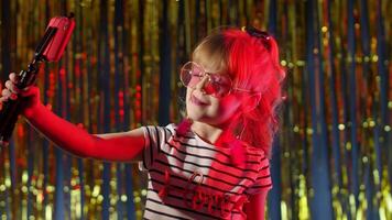 Fashion girl in futuristic night club with red neon lights making selfie call on mobile phone. Trendy stylish child kid blogger smiling cheerfully, recording blog vlog on smartphone with tripod video