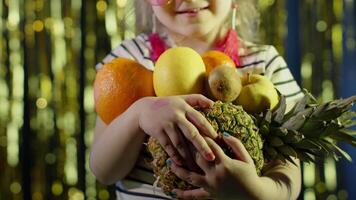 Close-up shot of girl in stylish sunglasses posing looking at camera with bunch of fruits in hands on shiny background in night club. Child kid sensual look clubber in neon yellow lights, slow motion video
