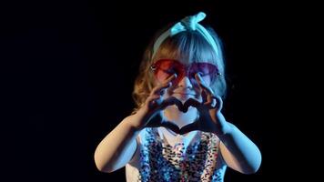Portrait of trendy stylish girl looking at camera, smiling, showing heart sign. Child kid in neon lighting posing at disco party cyberpunk studio. Futuristic hipster teenager children fooling around video