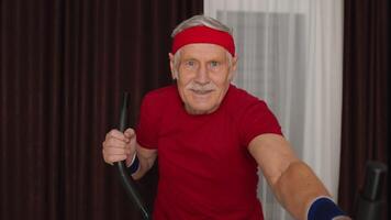 Old grey-haired senior grandfather model exercising workout on orbitrek in room at home. Retro style Caucasian mature elderly man warming up while sitting on orbitrek indoors. Healthy sport lifestyle video
