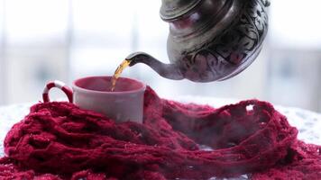 Pouring hot tea from ornate metal teapot into mug wrapped in red scarf and steam rise from cup in snow. Hand use long sleeve to hold hot kettle. Arabic style vintage teapot with traditional patterns video
