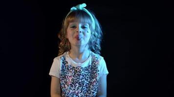 Portrait of trendy stylish girl showing tongue, fooling around. Elegant child kid in neon lighting posing at disco party cyberpunk studio. Futuristic hipster teenager children making faces slow motion video