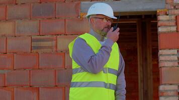 Professional builder engineer architect operate with radio walkie-talkie talking to control working at construction site. Foreman worker wearing hard hat, vest at building house. Business, real estate video