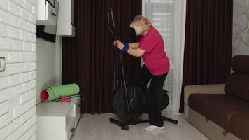 Senior elderly caucasian woman in sportswear using orbitrek in room at home doing sport training warming up cardio exercises. Active old grandmother during coronavirus lockdown. Healthy lifestyle video