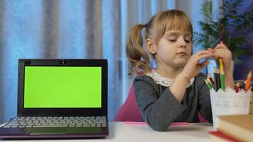 Child pupil schoolgirl at home sitting at table with digital laptop computer with green screen chroma key. Portrait of a kid girl distance education. Studying on coronavirus quarantine. Slow motion video