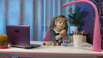 Child schoolgirl enthusiastically drawing with brushes and paints at home sitting at table using laptop digital computer. Portrait of kid girl distance education. Studying during coronavirus lockdown video