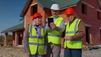 Diverse team of specialists taking near modern house. Real estate building project with senior civil engineer, architect, general worker discussing planning, development details on digital tablet video
