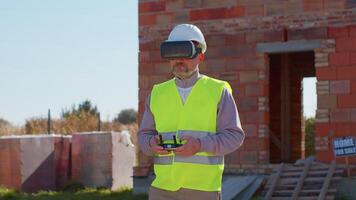 Drone operator controls an aircraft, holding remote controller. Professional builder in virtual reality helmet standing in front of construction site. House building. Business, real estate, investment video