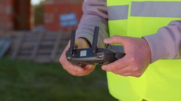 Drone operator controls an aircraft, hands holding remote controller close-up. Professional builder in vest standing in front of construction site. House building. Business, real estate and investment video