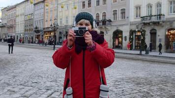 retrato de mayor mujer turista tomando imágenes con foto cámara, mirando a cámara utilizando retro dispositivo en invierno ciudad centrar de lviv, Ucrania. fotografía, de viaje, vacaciones. activo pensionista video
