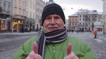Portrait of stylish senior man tourist smiling, showing thumbs up, looking at camera in winter city center of Lviv, Ukraine. Photography, travelling, vacation. Active pensioners life after retirement video