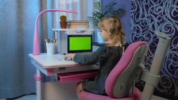 Child schoolgirl learns lessons being at home sitting at table using digital laptop computer with green screen chroma key. Portrait of a kid girl distance education. Studying on coronavirus quarantine video