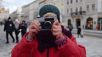 retrato de antiguo mayor mujer turista tomando imágenes con foto cámara, mirando a cámara utilizando retro dispositivo en invierno ciudad centrar de lviv, Ucrania. fotografía, de viaje, vacaciones. activo pensionista video