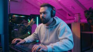 Addicted Caucasian guy male gamer streamer man play online battle enjoying smiling wearing headphones gaming at computer streaming in neon dark room at home match digital playing looking internet game video