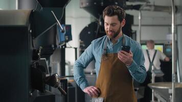 Caucasian man working in roasted coffee fabric factory inspecting roasting machine caffeine beans checking quality holding mobile phone texting typing smartphone smiling happy technologist male job video