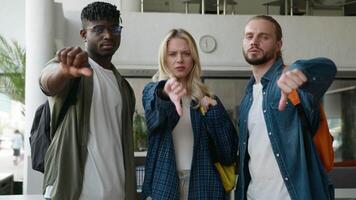 Three multiracial friends diversity students showing thumbs down negative dissatisfied bad dislikes looking at camera in university corridor hall together studying education problem unwell attitude video
