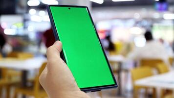 Smartphone with green screen held in hand at food court, Restaurant in a shopping mall, Blurred background with people. Perfect for mobile app or website content placement video