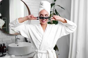 Woman in a spa robe enjoying a skincare routine while smiling in a modern bathroom with a towel on her head photo