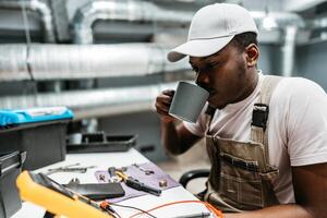Young man enjoying a coffee break while working on electrical equipment in a modern workshop setting photo
