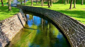 A serene park with a stone wall encompassing a river and pond. A charming bridge arches over the river, showcasing lush trees in the backdrop photo