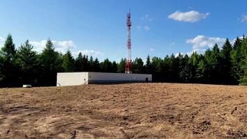A telecommunications tower with a facility situated in a cleared area surrounded by trees and under blue skies photo