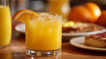 A refreshing drink filled with orange juice and ice sits on a wooden table. Behind it, plates of breakfast items are visible, highlighting a sunny summer morning. photo