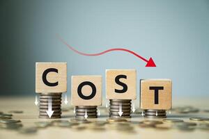 Concept of cost reduction in business finance with wooden blocks spelling COST placed on descending stacks of coins, red downward arrow, symbolizing decreasing expenses, saving strategy, efficiency photo