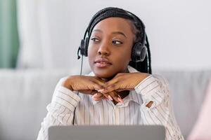Pensive young black lady having online training at home, sitting at workdesk in front of laptop, leaning on her elbows and looking at copy space, using headphones. E-education, online class concept photo