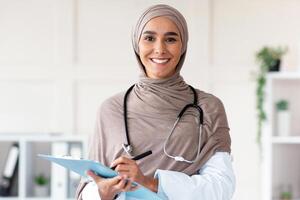 Cheerful pretty woman in hijab doctor posing in cabinet, holding medical folder and pen, having stethoscope, copy space. Happy muslim lady greeting patients at hospital, smiling at camera photo