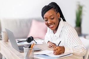 Pretty black woman student studying in front of laptop, taking notes and smiling, side view, copy space. Cheerful african american young lady attending webinar while staying home during lockdown photo
