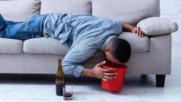 Alcohol Poisoning. Drunk Man Vomiting Into A Bucket After Drinking Alcoholic Wine Lying On Sofa At Home. Booze Intoxication, Alcoholism Addiction Habit Concept. photo