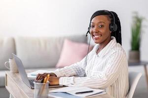 Pretty black woman smiling at camera while listening to webinar from home, using headset and laptop, side view, copy space. Cheerful african american lady working online, typing on computer keyboard photo