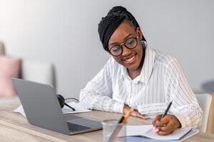 Cheerful african american business lady in formal outfit and eyeglasses attending webinar or business training, sitting at workdesk, using laptop at home, taking notes, copy space photo