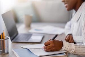 Closeup of young black woman having online course, smiling african american lady in headset sitting at table in front of laptop, taking notes in notepad, home interior, side view, copy space photo