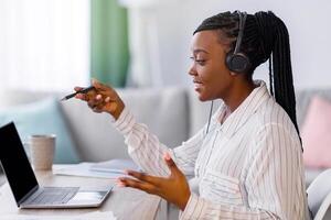 Beautiful young black woman freelancer with headset having chat from home, using laptop, looking at computer screen, talking and gesturing, side view, copy space. Working from home concept photo