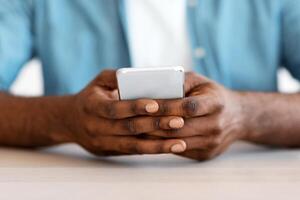 Closeup Shot Of African American Man Sitting At Desk And Using Smartphone, Cropped Image Of Unrecognizable Black Guy With Mobile Phone In Hands Texting With Somebody Or Browsing New App photo