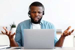 Working Stress. Annoyed Black Man In Headset Looking At Laptop Screen, Angry Call Center Operator Having Problems While Consulting Client, Sitting At Workplace With Computer, Free Space photo