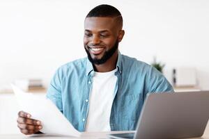 Millennial Black Freelancer Man Working With Papers At Desk In Home Office. Smiling African American Guy Sitting At Table With Computer Reading Documents And Checking Financial Reports, Free Space photo