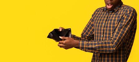 Shocked Black Guy Showing Empty Wallet At Camera, Suffering From Poverty And Absence Of Money, Emotional African Man Having Financial Crisis, Standing Isolated On Yellow Studio Background, Copy Space photo
