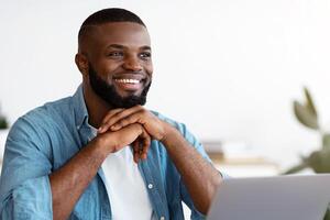 Portrait of pensive smiling black freelancer sitting at desk in home office, thinking about business strategy while working on laptop computer, looking away with pleased face expression, free space photo