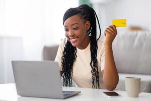 Cheerful young black woman with credit card using laptop at home, order food online, copy space. Smiling african american lady shopping or banking online, using computer, living room interior photo
